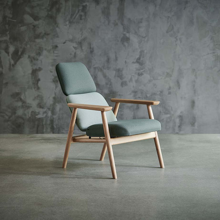 Lænestol, model Bear Recliner, med stel i massiv eg og polster i to nuancer grøn tekstil, stående foran betonspartlet væg